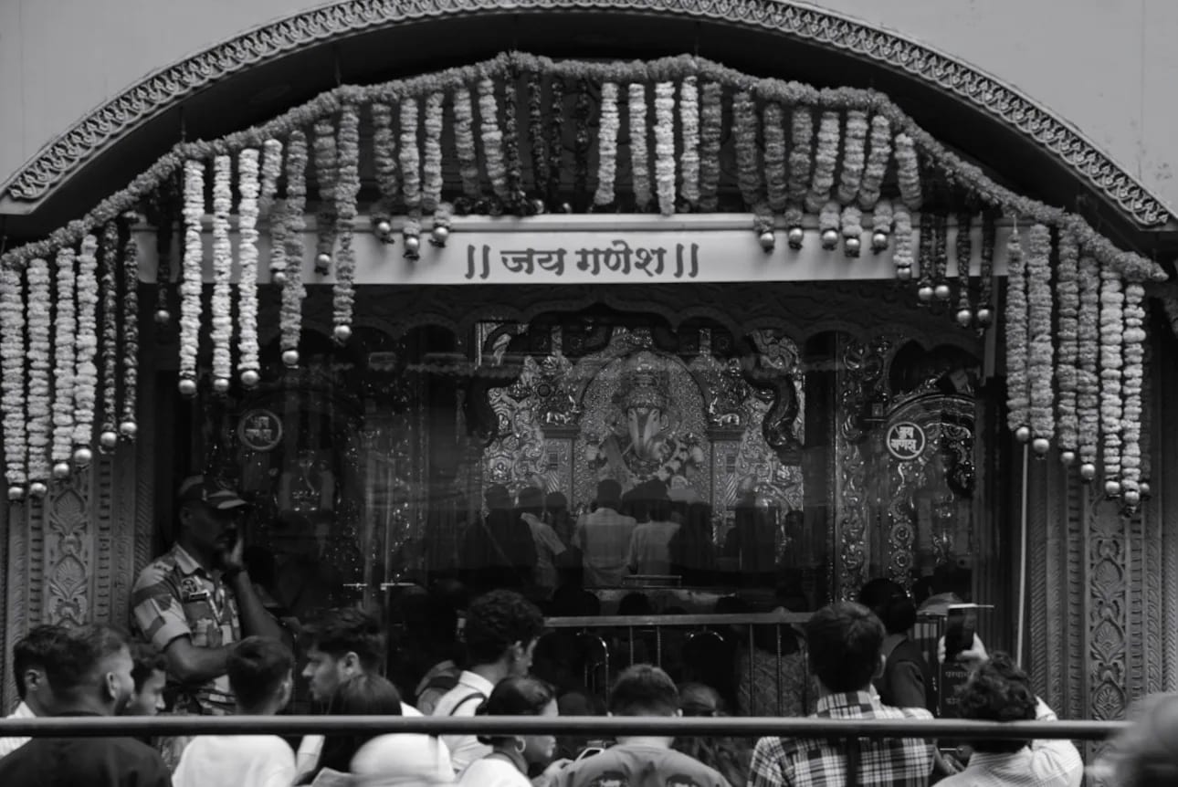 A decorated Ganpati pandal hung with garlands, a crowd seated in front of it, in black and white