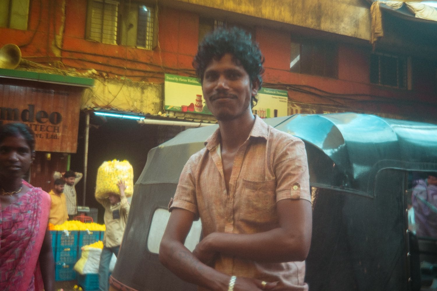 A man standing beside his autorickshaw in a market lane, stalls and produce behind him