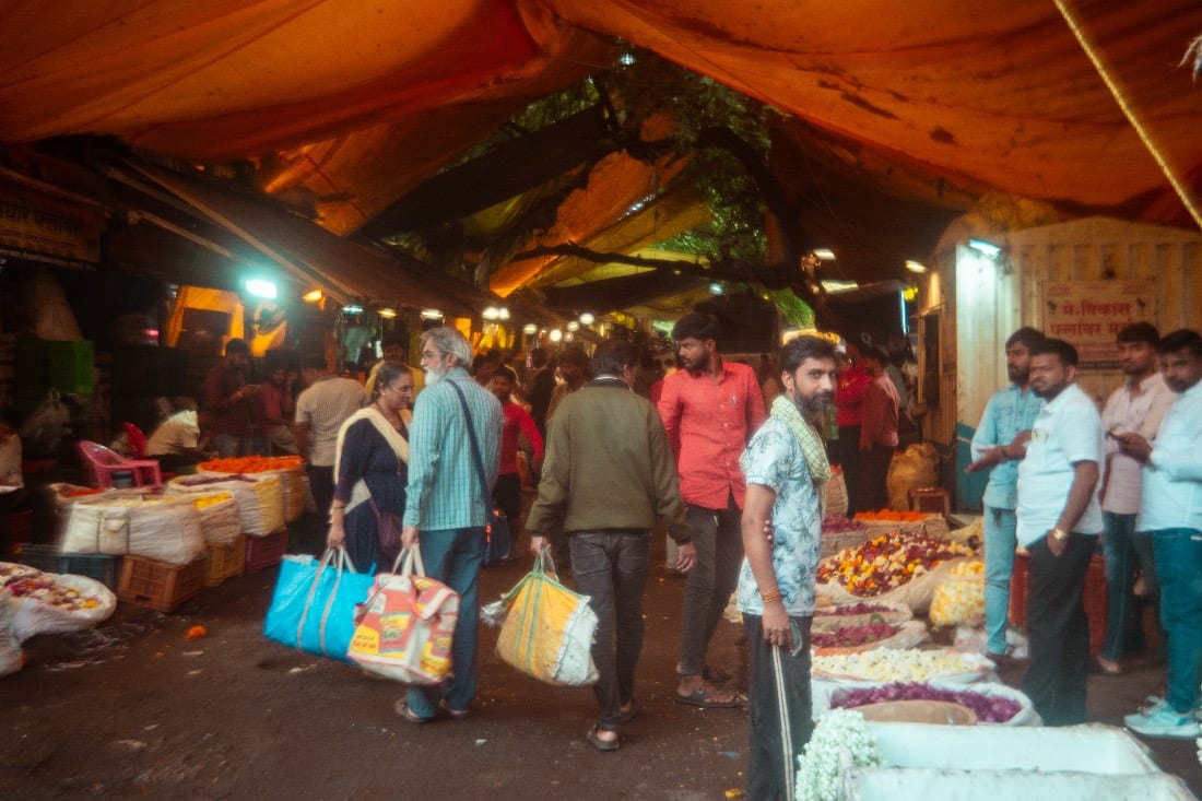 A crowded market under an orange canopy, shoppers walking between stalls laid out with flowers