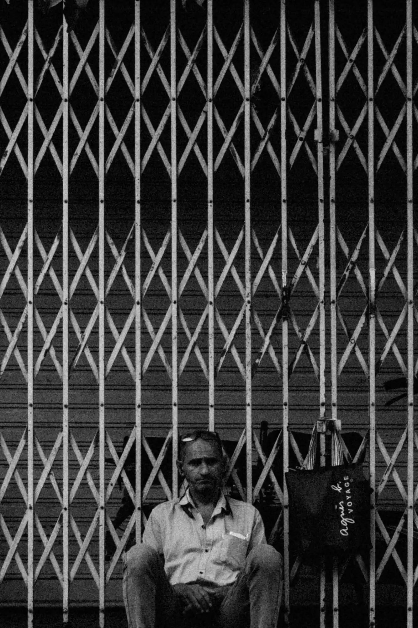 A man sitting behind a folding metal gate, its lattice filling the frame, in black and white