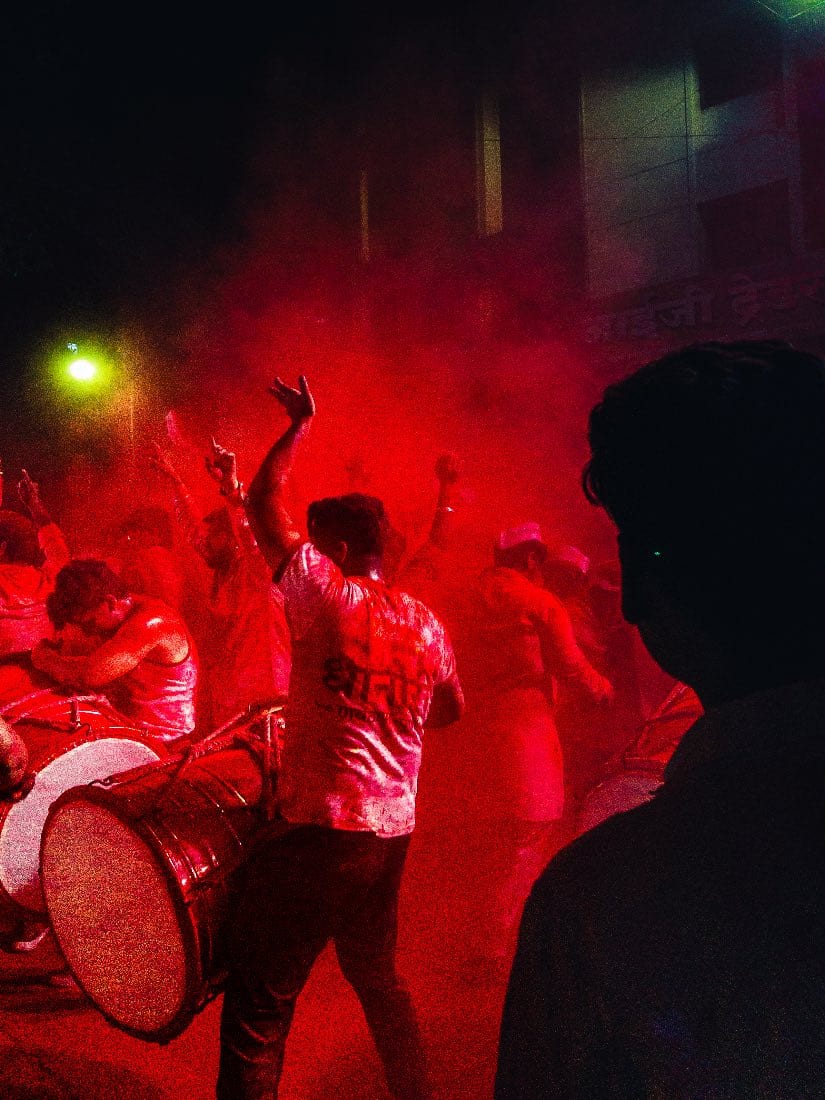 A drummer in a crowd lit red at night, smoke and raised arms around him