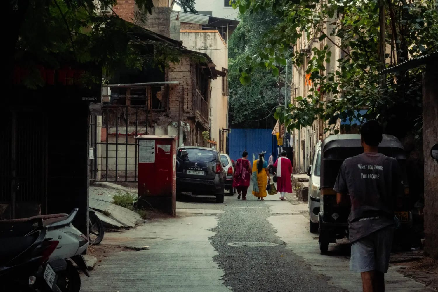 A narrow Pune lane with people walking away between old buildings, an autorickshaw parked to one side and greenery overhead