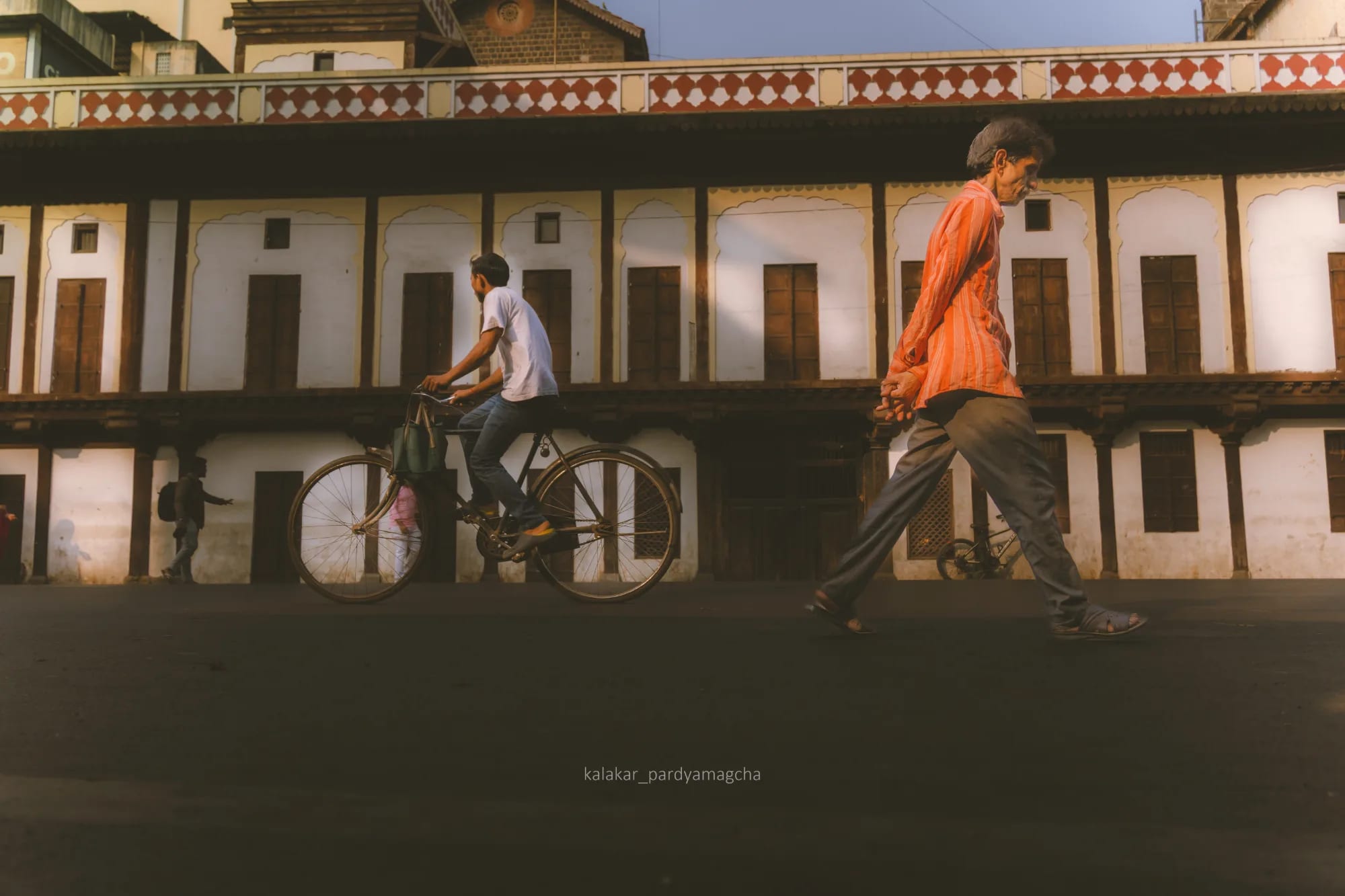 A cyclist and a man in orange passing the long arched facade of an old wada
