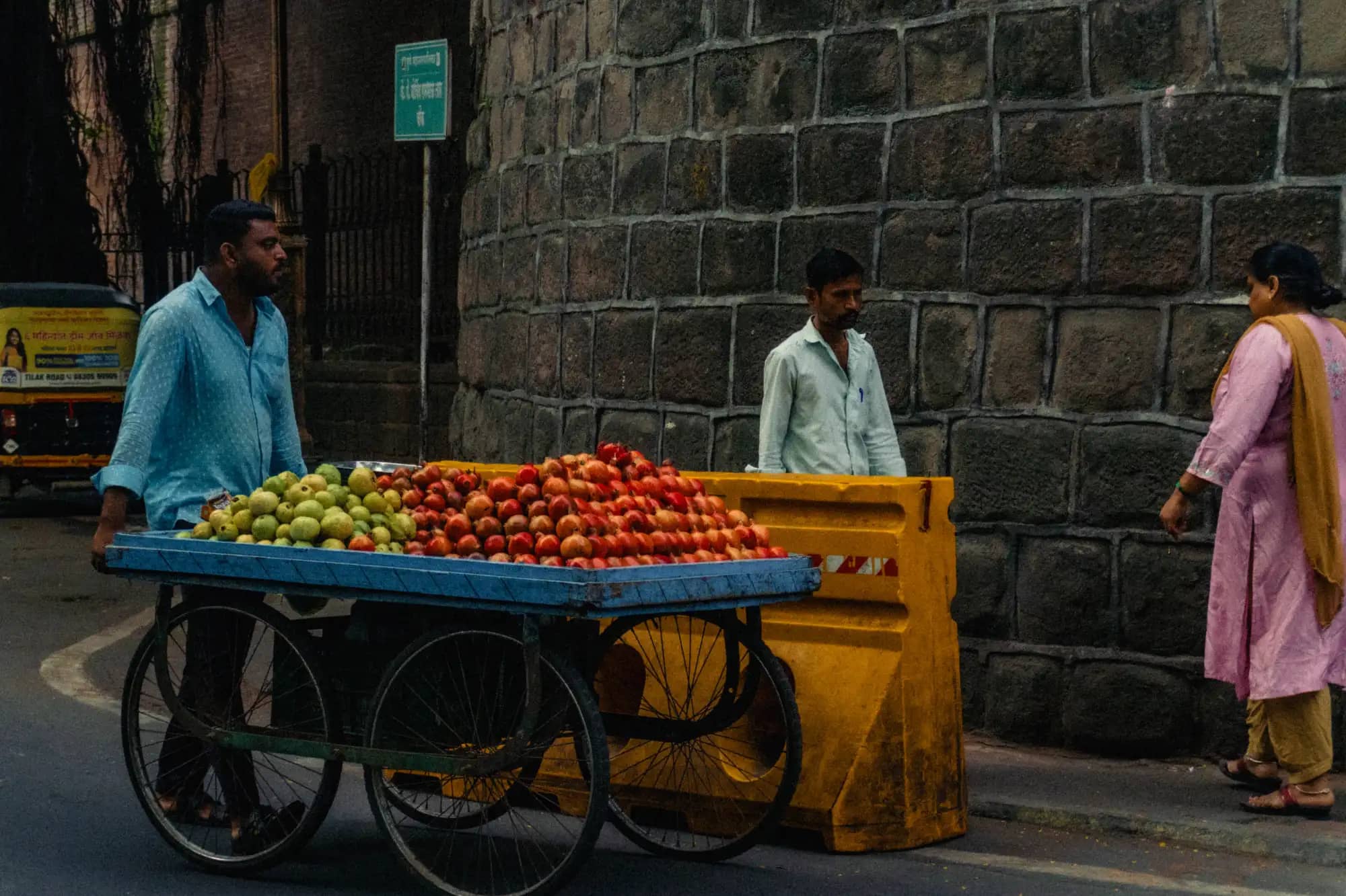 A handcart of tomatoes and limes against a stone wall, its seller waiting beside it