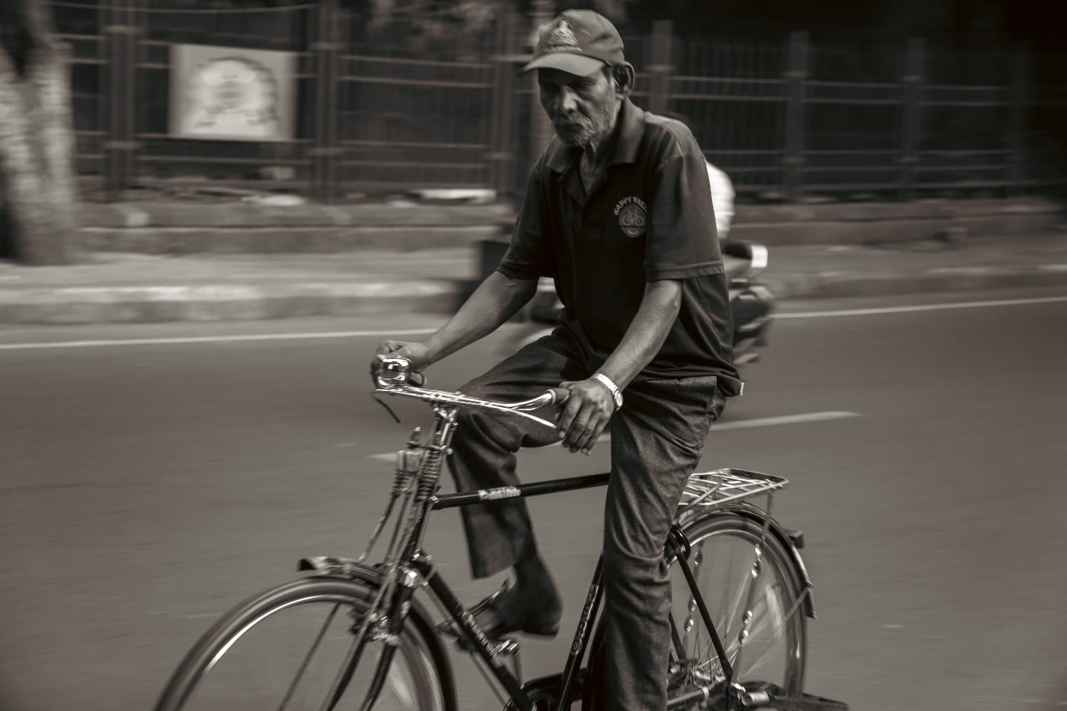 An older man riding a bicycle, the road behind him blurred with movement, in black and white